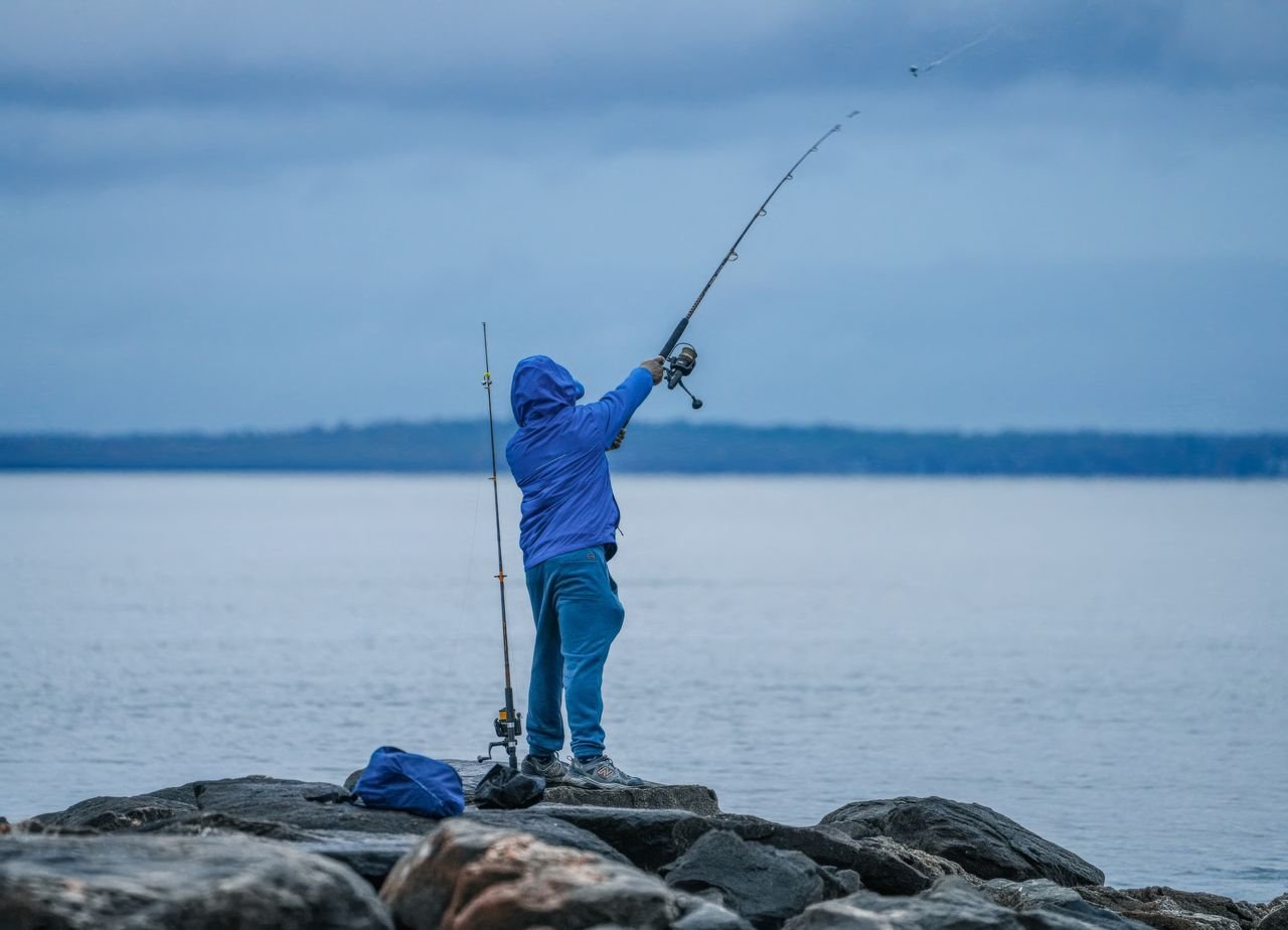 Fishing A fisherman in blue clothing casts a line off rocky shores into a calm lake under a cloudy sky.