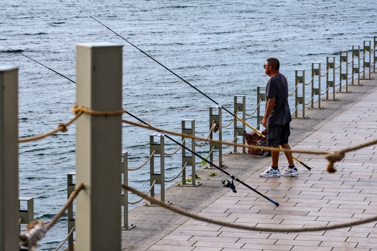 Resources A man stands fishing with rods along a waterfront promenade, enjoying the peaceful day.